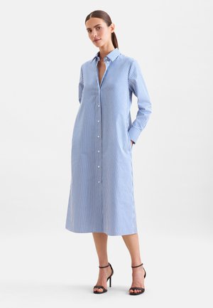Une femme porte une robe chemise midi à rayures bleues et blanches avec boutons sur le devant, accompagnée de sandales noires à talons hauts, sur un fond blanc uni.