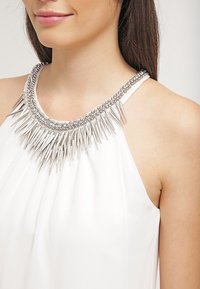 Vit halterneck-topp med en texturerad halsringning prydd med silverpiggar och pärlor, vilket skapar ett distinkt mönster och kontrast.