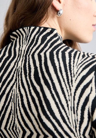 Μαύρο και κρεμ ριγωτό ρούχο με ψηλό λαιμό και υφή με κυματιστό μοτίβο. Ασημένιο κυκλικό σκουλαρίκι στο αυτί του μοντέλου.