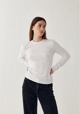 Weißes Langarmshirt mit Rundhalsausschnitt, weichem Material und figurbetontem Schnitt. Kombiniert mit schwarzen Cordhose mit vertikalen Streifen.