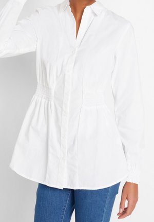 Donna che indossa una camicetta bianca a maniche lunghe con bottoni, vita e maniche arricciate, abbinata a jeans blu.