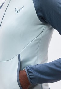 Persona que lleva una chaqueta de manga larga de color gris claro y azul oscuro con una mano en el bolsillo delantero y un pequeño logo de animal en el pecho.