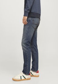 Jeans de denim azul con un diseño de pierna recta, lavado medio y un sutil desvanecimiento; combinados con zapatillas blancas con acentos verdes y rojos.