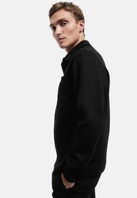 Svart sweatshirt med hög krage, tillverkad av ett texturerat tyg, med en sidoficka och kontrasterande sömnadsdetaljer. Tätt passform.