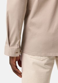 Camicia beige chiaro a maniche lunghe con polsini abbottonati, texture liscia e orlo arrotondato, abbinata a pantaloni avorio con tasca sul retro.