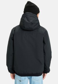 Personne portant une veste à capuche noire et un bonnet noir, de dos, associée à un jean en denim bleu clair sur un fond uni.