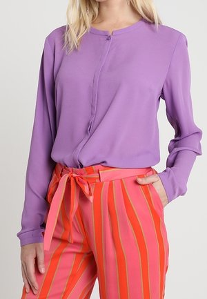 Vrouw in een paarse blouse met lange mouwen, ingestopt in een roze broek met hoge taille en oranje verticale strepen, afgewerkt met een ceintuur die is gestrikt.