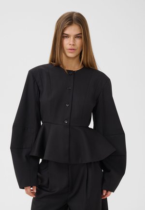 Femme aux cheveux longs et raides portant une veste péplum noire boutonnée avec de larges manches et un pantalon noir assorti sur un fond clair.
