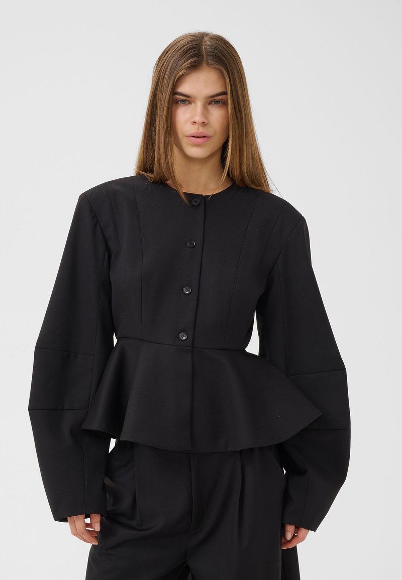 Femme aux cheveux longs et raides portant une veste péplum noire boutonnée avec de larges manches et un pantalon noir assorti sur un fond clair.