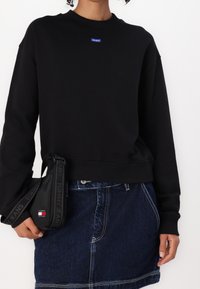 Schwarzes Sweatshirt mit Rundhalsausschnitt und langen Ärmeln, ausgestattet mit einem blauen Logo-Patch. Kombiniert mit einem Jeansrock und einem schwarzen Tasche-Riemen.