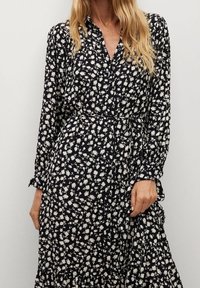 Schwarzes Kleid mit weißem Blumenmuster, langen Ärmeln, geknöpftem Kragen und tailliertem Schnitt. Fließendes Design mit schichteter Rock. Weicher Stoff.