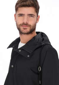 Schwarze wasserdichte Jacke mit Kapuze, ausgestattet mit Druckknöpfen, Reißverschluss und Kordelzug-Details. Glatter Stoff, entworfen für Komfort und Funktionalität.
