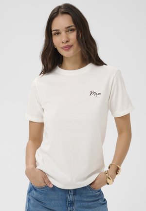 Jeune femme aux cheveux foncés portant un T-shirt blanc avec le texte « Mom », jean bleu et bracelets dorés, mains dans les poches, fond neutre.