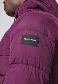 Fioletowa kurtka puchowa o fakturowanym wykończeniu, z czarną prostokątną naszywką z logo na lewym ramieniu, lekka i ocieplana.