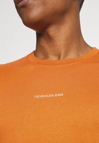 Oranžové bavlněné tričko s kulatým výstřihem a krátkými rukávy, na přední straně s bílým potiskem "CALVIN KLEIN JEANS".