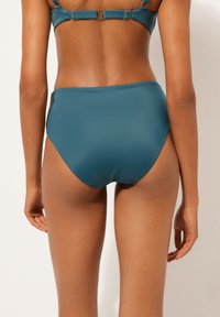 Högmidjad bikinitopp i teal-tyg. Har en mjuk textur med en ren skärning och subtila sidodetaljer. Bakifrån visar passform och täckning.