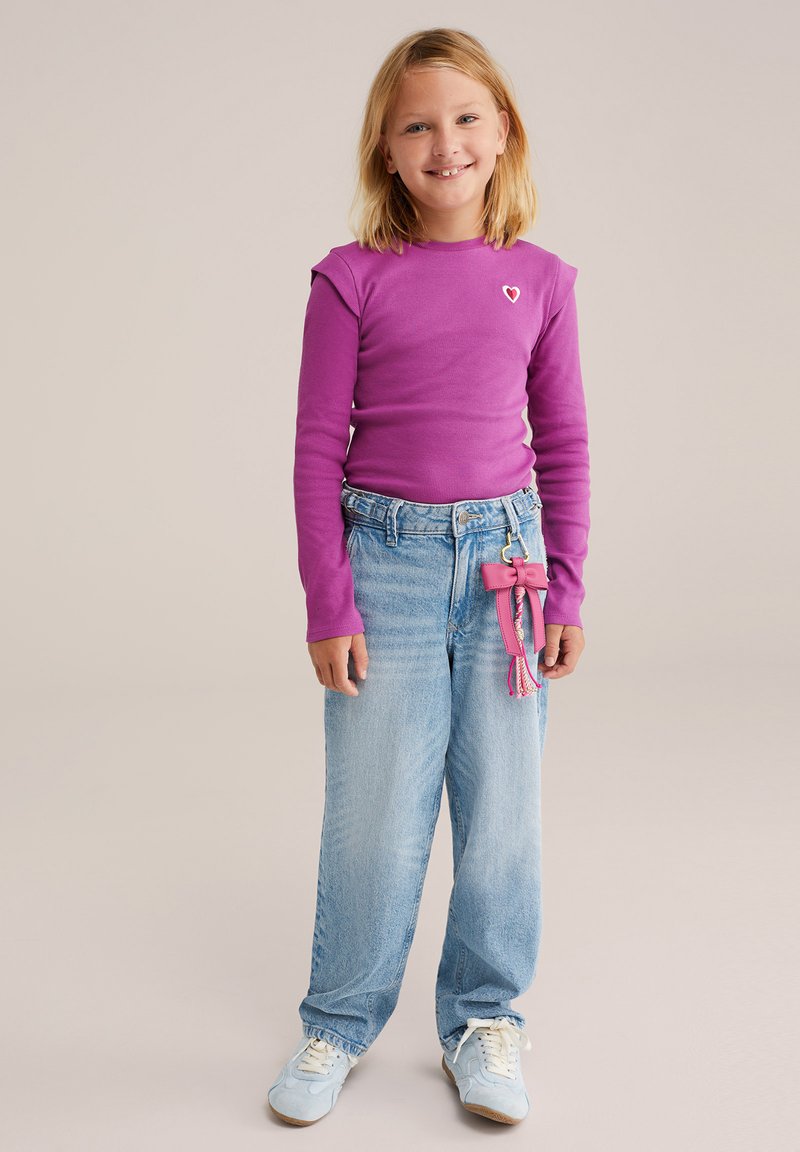Haut violet à manches longues avec des détails aux épaules, un pantalon en denim bleu et des baskets bleu clair. Porte-clés rose avec des pompons attaché au pantalon.