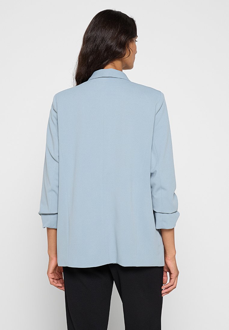 Blazer bleu clair à coupe décontractée, col à cran et manches retroussées. Tissu doux avec une texture lisse, sans motifs visibles ni matériaux métalliques.