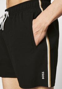 Shorts de bain noir avec une taille élastique, cordon blanc, poches latérales et bandes latérales dorées et blanches. Caractérisé par le logo BOSS au bas.
