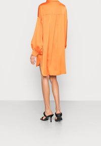 Vestido camisolín de satén naranja con mangas largas y abullonadas, detalle de pliegue en la espalda, aberturas laterales y combinado con sandalias de tacón negras.