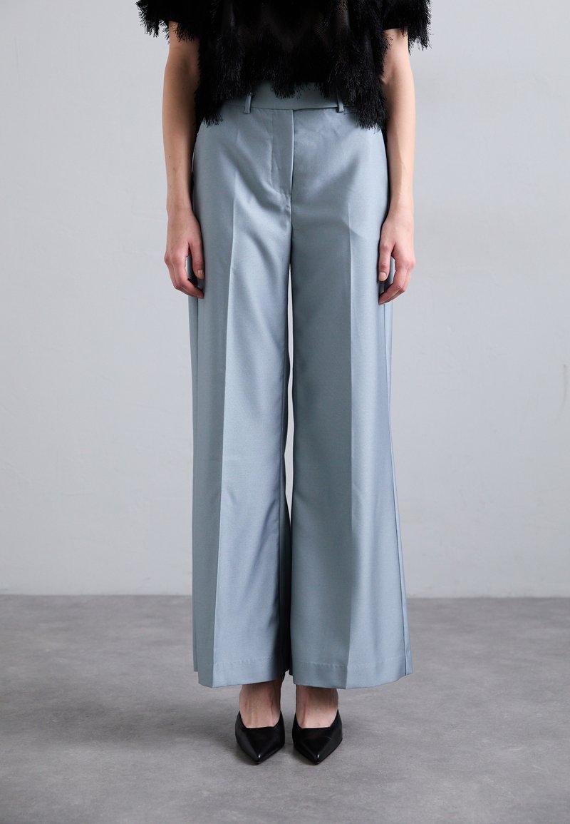 Jasnoniebieskie szerokie spodnie wykonane z gładkiego materiału, z dopasowanym fasonem w talii i ostrymi kantami, w połączeniu z czarnymi szpilkami.