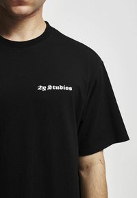 Juodas medvilnės marškinėliai su baltu "2y Studios" logotipu kairėje krūtinės pusėje. Trumpomis rankovėmis ir standartinio dėvėjimo stiliumi. Lygi tekstūra.
