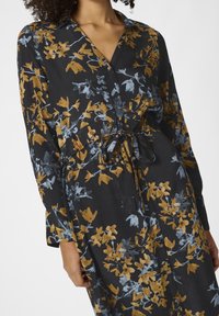 Femme portant une robe noire à manches longues avec un motif floral bleu et jaune moutarde et une ceinture nouée à la taille.