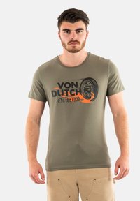 Camiseta de algodón verde oliva con un gráfico negro y naranja que presenta "VON DUTCH" y un diseño de neumático. Mangas cortas y cuello redondo.