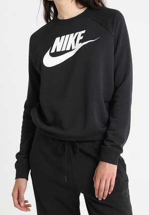 Vrouw draagt een zwarte Nike-trui met wit logo en bijpassende zwarte joggingbroek, staand tegen een effen achtergrond.