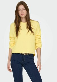 Pull en maille jaune avec un col arrondi et des poignets côtelés, associé à un jean bleu foncé ajusté et une ceinture avec des accents dorés.