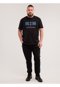 Habeme jaaga mees, kes kannab musta T-särki, millel kiri "BIG STAR", musti pükse ja musti tossusid, seisab ühevärvilise heleda tausta ees.