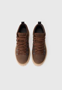 Pantofi de drumeție din suede maro, cu șireturi portocaliu-maroniu, ochiuri metalice, tălpi bej și bucle negre pentru călcâi, văzuți din sus pe un fundal alb.
