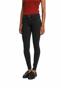 Femme portant un jean skinny noir, des mocassins noirs et un haut côtelé rouge sans manches, debout, les bras détendus le long du corps.