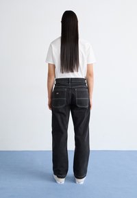Pantaloni din denim negri cu cusături albe și două buzunare spate. Asociați cu un tricou simplu alb. Modelul se întoarce, evidențiind designul.