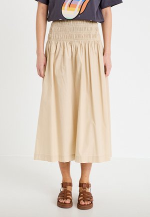 Beżowa spódnica midi z szerokim marszczonym pasem, noszona z czarnym t-shirtem z grafiką i brązowymi sandałami na cienkich paskach na stojącej osobie.