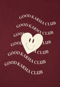 Bordo majica s kratkimi rokavi, ki ima bel srček s nasmejanim obrazom, obdan s ponavljajočim se besedilom "DOBRA KARMA KLUB" v belem, valovitem fontu.