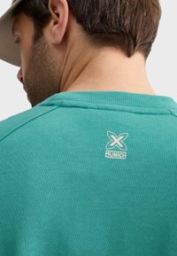 Tyrkysový sveter s rebrovaným golierom a malým bielym logom "X MUNICH" na chrbte. Mäkká textúra, voľnočasový dizajn.