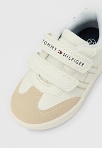 Scarpa da ginnastica beige e bianca per bambini con due strap in Velcro, marchiata "Tommy Hilfiger" con logo sulla fascia superiore.