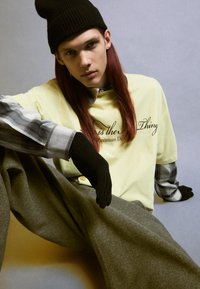 Tricou galben supradimensionat cu text negru, cămașă cu mânecă lungă în carouri gri dedesubt, mănuși negre și pantaloni largi gri. Modelul stă sprijinit de un fundal gri.