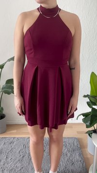 Žena v bezrukávových burgundských halenkách s halter krkom stojí v interiéri pri zelených kvetináčoch na svetlej drevenej podlahe a šedom koberci.