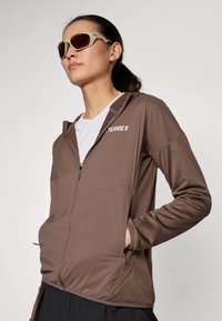 Giacca marrone con zip, maniche lunghe e cappuccio testurizzato, dotata di un logo bianco. Completata da occhiali da sole con montatura chiara e lenti marroni.