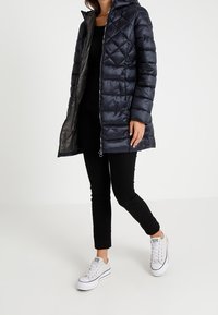 Navy blue quilted puffer coat with a hood, front zipper, and two side pockets, worn over a black top and black pants, paired with white sneakers.
