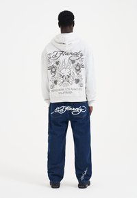 Grå hættebluse med ryggrafik af et kranium og flammer. Sammen med løse blå jeans med hvidt mærke-logo på lommerne.