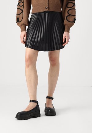 Černá plisovaná sukně spárovaná s hnědým cardiganem, který má černé křivolaké výšivky. Model má na sobě robustní černé boty na kotník.