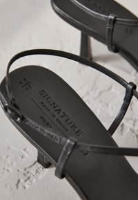 Sandales à talons en cuir noir avec des lanières fines et une semelle intérieure texturée. Présente l'inscription "SIGNATURE MADE IN BRAZIL" embossée sur la semelle.