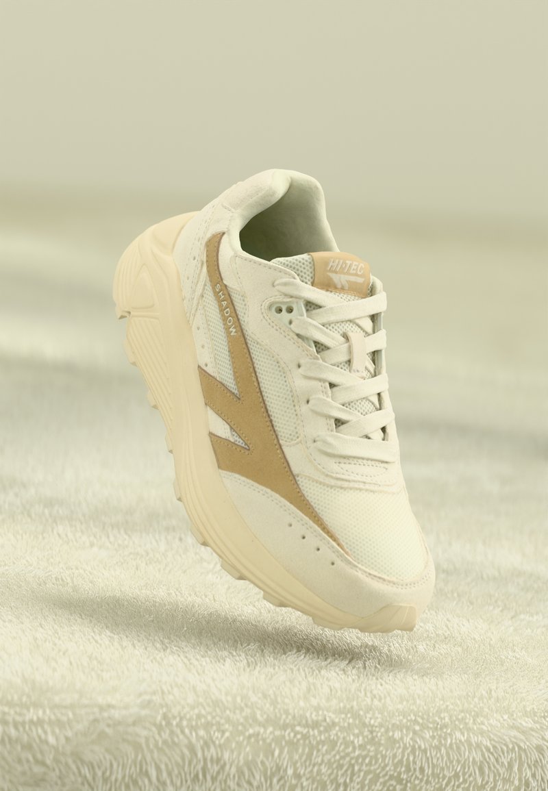 Adidași albi cu accente bej deschis, materiale din plasă și piele suferință, vârf rotund, sistem de prindere cu șireturi și talpă texturată. Numele brandului "HI-TEC" vizibil.