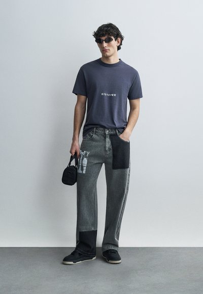 Γκρι υπερμεγέθη μπλούζα με το λογότυπο "KID SUPER" τυπωμένο σε λευκό. Γκρι τζιν με πατς και ανάμεικτες υφές. Μαύρα αθλητικά παπούτσια και μικρή τσάντα. Γυαλιά ηλίου.