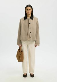 Femme portant une veste beige à col noir, un pull beige, un pantalon blanc, des chaussures marron, tenant un sac fourre-tout en daim marron, debout devant un fond blanc.