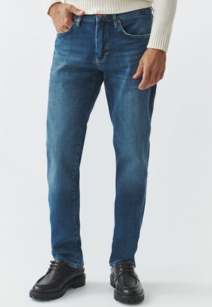 Jeans de mezclilla azul oscuro con corte recto, ligero desgaste, cinco bolsillos y costuras en contraste. Llevados con zapatos negros de cordones.