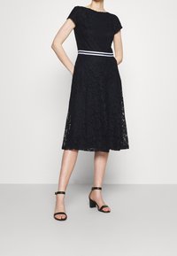 Žena pózuje v černých krajkových šatech ke kolenům s páskem se bílými pruhy a černými sandály na blokovém podpatku s kotníkovým páskem na jednobarevném pozadí.